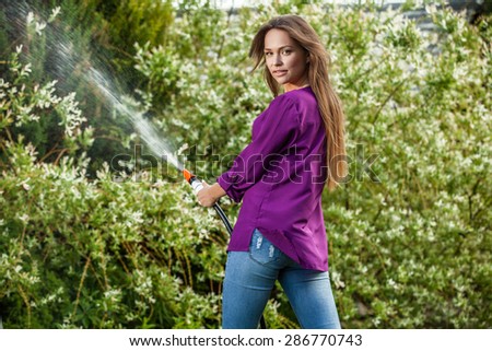 Beautiful joyful young girl in violet shirt poses in a summer garden with a water hose.
