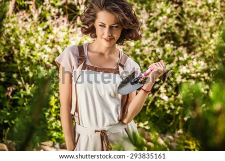 Outdoors portrait of beautiful & positive young woman in overalls which posing with mini colorful shovel in solar summer garden.