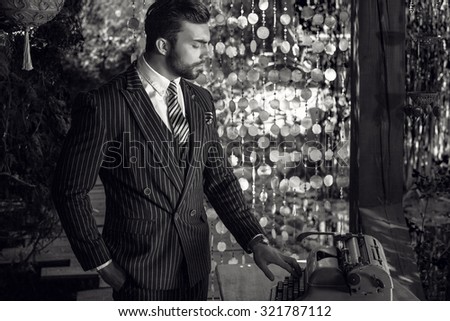 Black-white portrait of young beautiful fashionable man in classic suit in summer lodge near old typewriter.