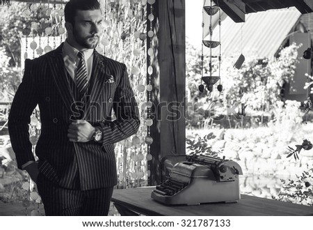 Black-white portrait of young beautiful fashionable man in classic suit in summer lodge near old typewriter.