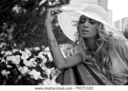 Portrait of beautiful blond woman in white retro hat siting near park flowers. Black-white photo.