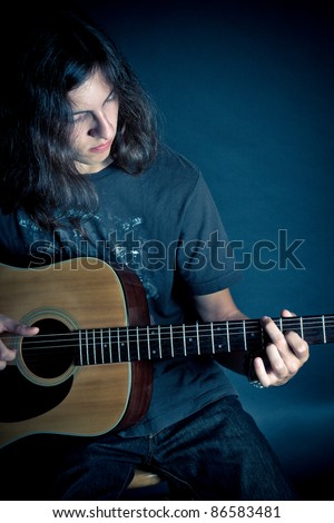 A young guy with long hair playing on an acoustic guitar against a dark background