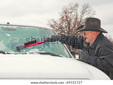 Man wearing a cowboy hat scraping ice off his vehicle windshield after an ice storm on a cold winter morning