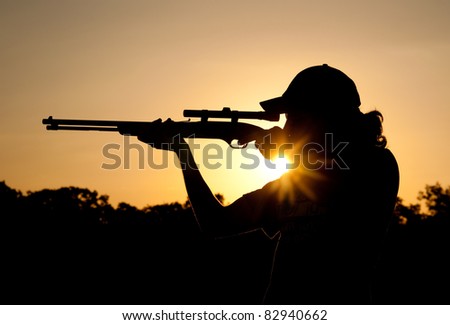 Silhouette of a young man shooting with a long rifle against sunset sky, with a sunburst