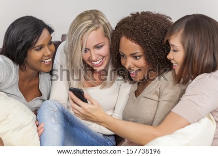 Interracial group of three beautiful young women girl friends at home sitting together on a sofa smiling, surprised and shocked using cell phone smartphone