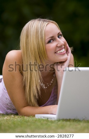 A beautiful young woman laying down in a grassy sunlit setting and working on her laptop