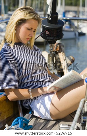 A beautiful young blond woman sitting on the deck of a sailing boat reading a book with her mobile phone on the deck beside her