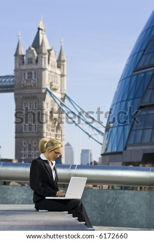 A beautiful young female executive working on her laptop in London, England, with her mobile phone beside her and Tower Bridge and Canary Wharf in the background