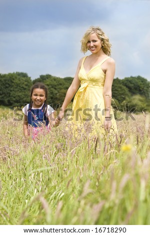 A beautiful blond haired blue eyed young woman having fun walking through a field with a mixed race young girl in a field of long grass