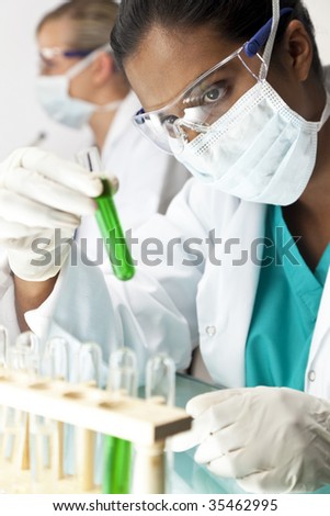 An Asian medical or scientific researcher or doctor using looking at a green solution in a laboratory with her female colleague out of focus behind her.
