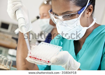 An Asian medical or scientific researcher or doctor working with a pipette, blood samples and a well tray  in a laboratory with her female colleague out of focus behind her.