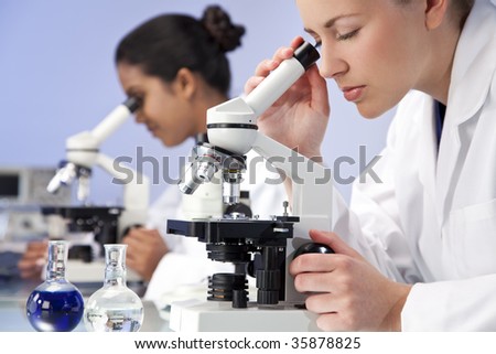 A blond female medical or scientific researcher or doctor using her microscope in a laboratory with her Asian colleague out of focus behind her.