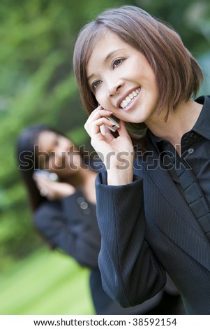 A beautiful young Chinese Asian oriental woman with a wonderful smile chatting on her cell phone, behind her is an Indian Asian colleague on her cell phone.