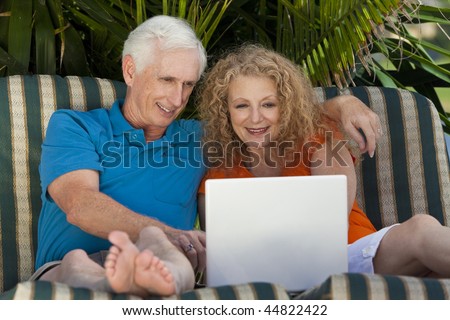 A senior retired man and woman couple sitting outside on sun loungers using a wireless laptop computer to surf the internet.
