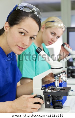 A female medical or scientific researcher or doctor using her microscope in a laboratory with her  colleague out of focus behind her.