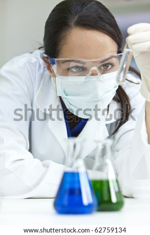 A female medical or scientific researcher or woman doctor looking at a test tube of clear solution in a laboratory with her conical flasks of green and blue solutions in front of her.