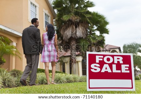 A happy African American man and woman couple house hunting outside a large house with a For Sale sign. The focus is on the sign.