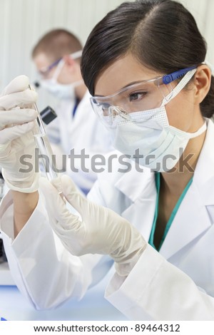 A Chinese Asian female medical or scientific researcher or doctor using looking at a test tube of clear liquid in a laboratory with her colleague out of focus behind her.
