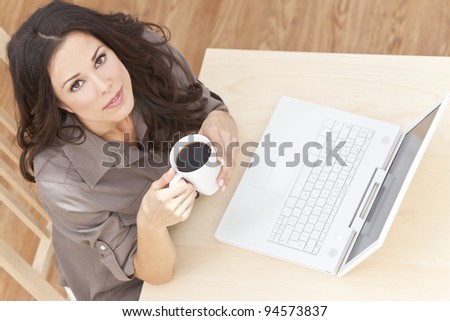 Beautiful, smiling, young brunette woman at home at a table using her laptop computer drinking a mug of tea or coffee