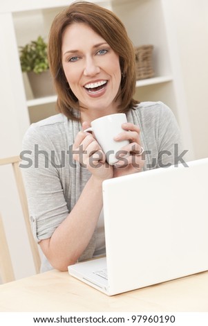 Beautiful, smiling, woman at home at a table using her laptop computer drinking a mug of tea or coffee