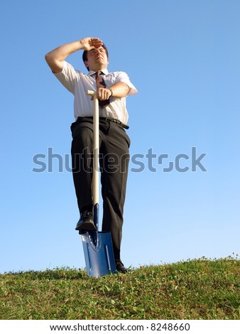 Businessman in white shirt and tie standing in grass field over clear blue sky with one leg resting on shovel driven into grass looking ahead - business vision concept