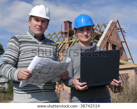 Female and male building engineers wearing helmets, standing against unfinished brick house, man holding building plans and woman laptop