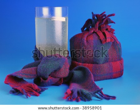 Effervescent tablet in glass of water wrapped around by scarf and cap next to it over blue background