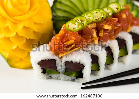 Fancy spicy tuna roll roll with avocado and jalapeno pepper on a white plate garnished with sliced cucumber and carved mango.
