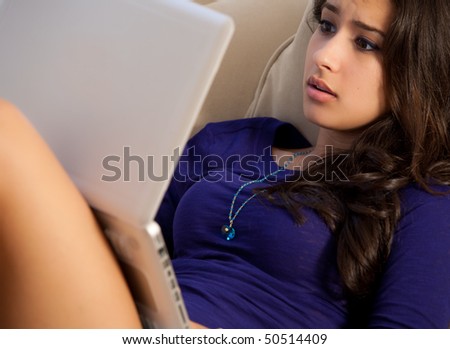 Beautiful young woman lying on a sofa surfing the internet on a laptop computer with a surprised expression.