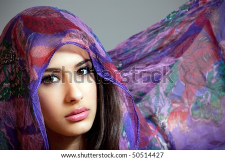 Beautiful young woman of multiple ethnicity in a glamor/fashion pose with a shawl partially covering her face on a gray background.