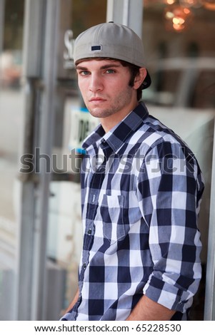 Handsome young man in an urban lifestyle fashion pose leaning against a store front wearing a baseball hat.