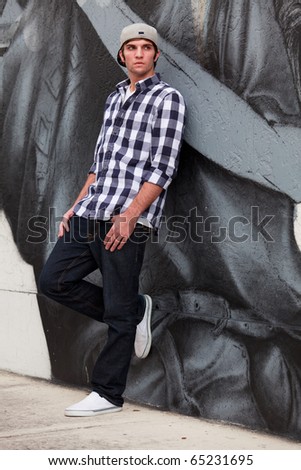 Handsome young man in an urban fashion lifestyle pose leaning on a graffiti wall wearing a baseball cap.