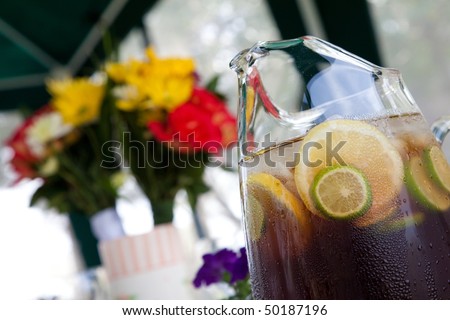 Sweet iced tea in a glass pitcher with lemon and lime fruit slices floating with a flower bouquet in the background