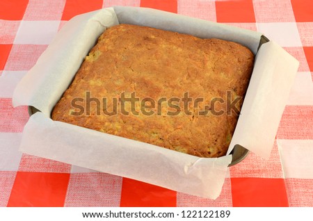 Parchment lined cake pan with cooked carrot cake cooling on red and white checked cloth