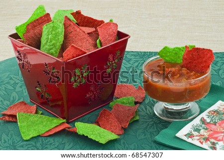 Red and green tortilla chips in christmas theme container with black bean and corn salsa on green mat