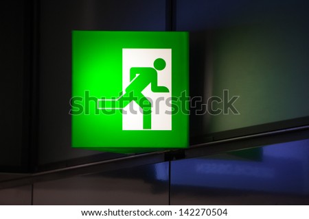 Illminated green exit sign attached to a wall in a public transportation facility. Signage consists of a human figure running and exiting through a door opening.