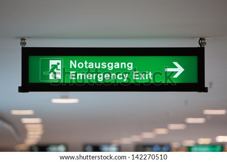 Illuminated green emergency exit sign hanging from the ceiling in a public transportation facility. Sign shows a figure exiting through a door and the word 