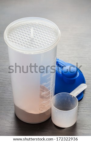 Chocolate whey isolate protein in the translucent protein shaker, next to the shaker parts and empty spoon