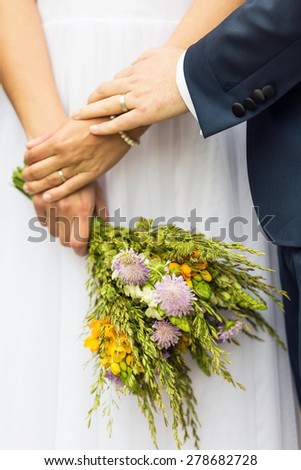 Bride holding wedding bouquet made of forest flowers in her hand, against the white lace of the wedding dress; groom holding the bride\'s hand