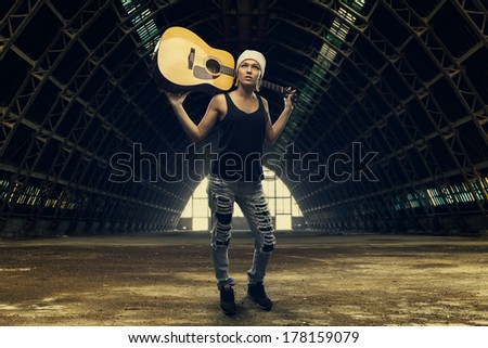 Young woman posing with an acoustic guitar in an abandoned factory hall. Toned image with low light effect.