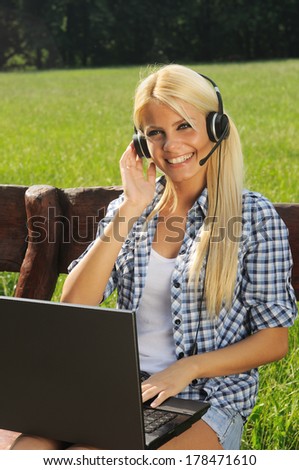 Female student sitting on wooden bench in the park with laptop in lap and headset