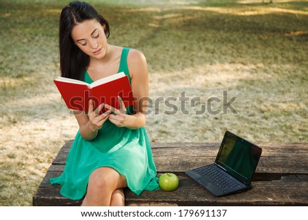 Beautiful young woman with toothy smile reading book in the park and sitting on a wooden table beside a green apple and laptop.