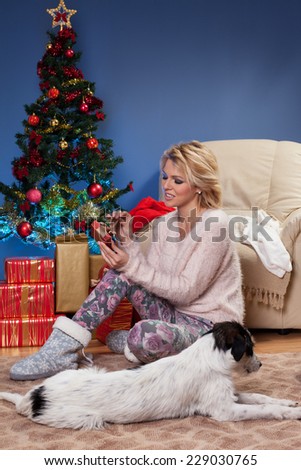 Beautiful young woman sitting near Christmas tree and opening a gift with the dog in front of her who sleeps on the floor.