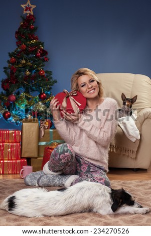 Beautiful young woman sitting near Christmas tree and holding a gift in arms with the dog in front of her who sleeps on the floor.