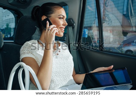 Young businesswoman talking on the phone in the back seat of the car and holding in hand a digital tablet