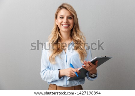 Photo of gorgeous blond secretary woman with long curly hair writing down notes in clipboard while working in office isolated over gray background