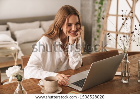 Photo of Image of young pleased happy cheerful cute beautiful business woman sit indoors in office using laptop computer listening music with earphones.
