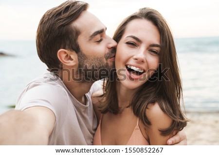Photo of Image of young happy man kissing and hugging beautiful woman while taking selfie photo on sunny beach