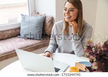 Photo of cheerful young woman in eyeglasses using laptop while having breakfast at living room