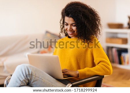Image of cheerful african american woman using laptop while sitting on chair in living room